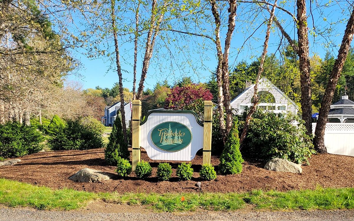 The Trailside Estates entrance sign in spring, with birch trees, mulched beds of boxwood and rhododendron, and a neighbor's home visible beyond at this 55+ active adult community in Rochester, MA