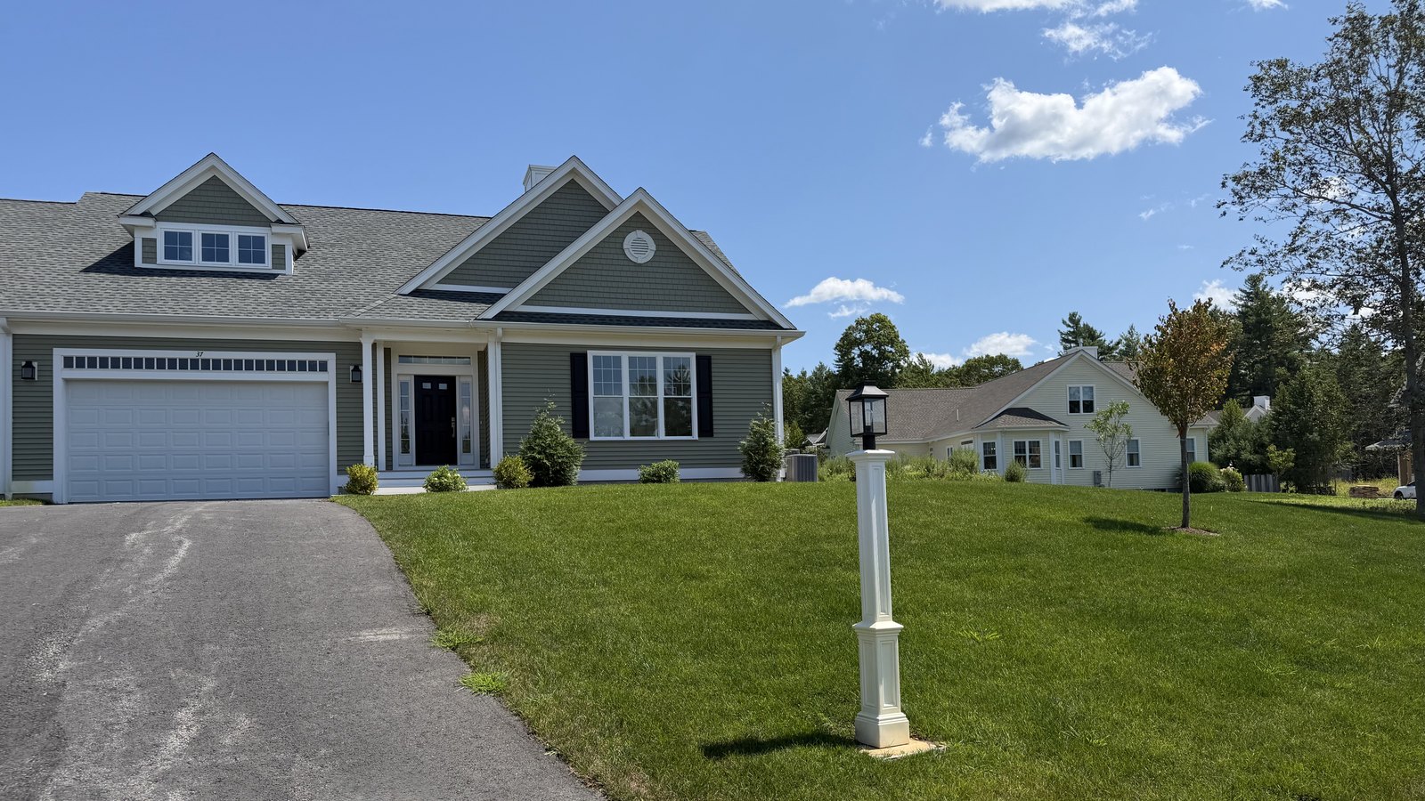 Front exterior of 37 Crestwood Circle at Trailside Estates, a 55+ active adult community in Rochester, MA — sage-green shingled duplex-style home with white trim, attached two-car garage and lamp post on a green lawn, open for touring Sunday, September 20